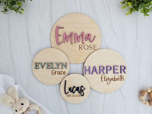 Flat lay of four personalized wooden baby name signs in various sizes, each featuring a 3D first name in colorful raised letters and an engraved middle name. Names shown are Emma Rose, Evelyn Grace, Harper Elizabeth, and Lucas Michael. The signs are placed on a white wood background with greenery and baby toys around the edges.